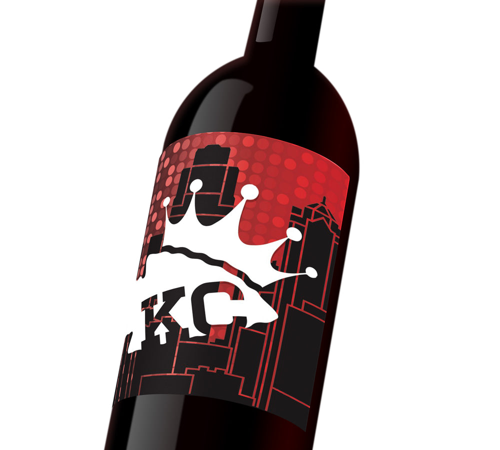 A red custom wine label with the KC skyline in the behind a white KC Chiefs spearhead that reads, "KC, wearing a KC Royals crown.