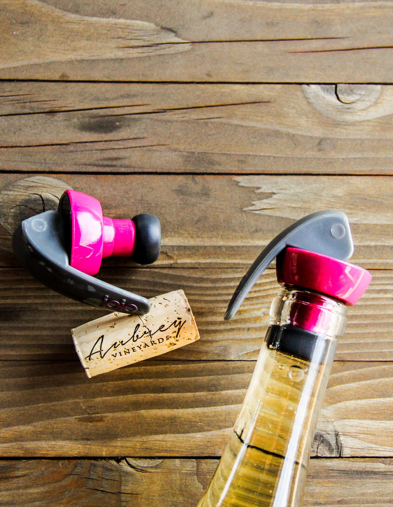 
                  
                    Load image into Gallery viewer, Two Joie bottle stoppers, one laying next to an Aubrey Vineyards cork and the other capping a bottle of Aubrey Vineyards wine,
                  
                