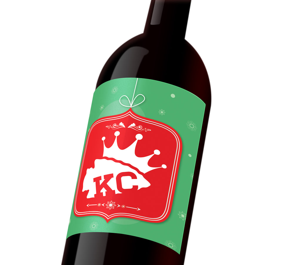 A green holiday custom wine label with a red holiday ornament depicting a white KC Chiefs spearhead wearing a KC Royals crown.